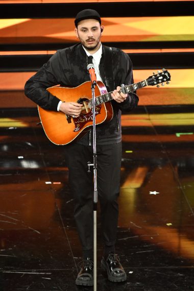 Il cantante delle nuove proposte indossa un bomber in nylon di seta nera con ricami macramè dai motivi floreali, pantalone jogging, camicia e cravatta black