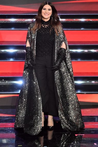 Lungo mantello luccicante su top traforato e pantaloni scuri per la cantante sul palco dell'Ariston
